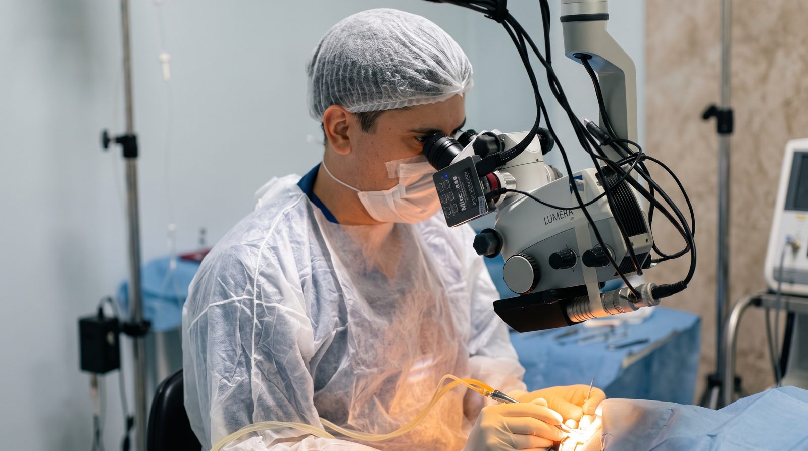 Dr. Marcus Felipe operando com microscópio cirúrgico, de touca e máscara, em sala de cirurgia.